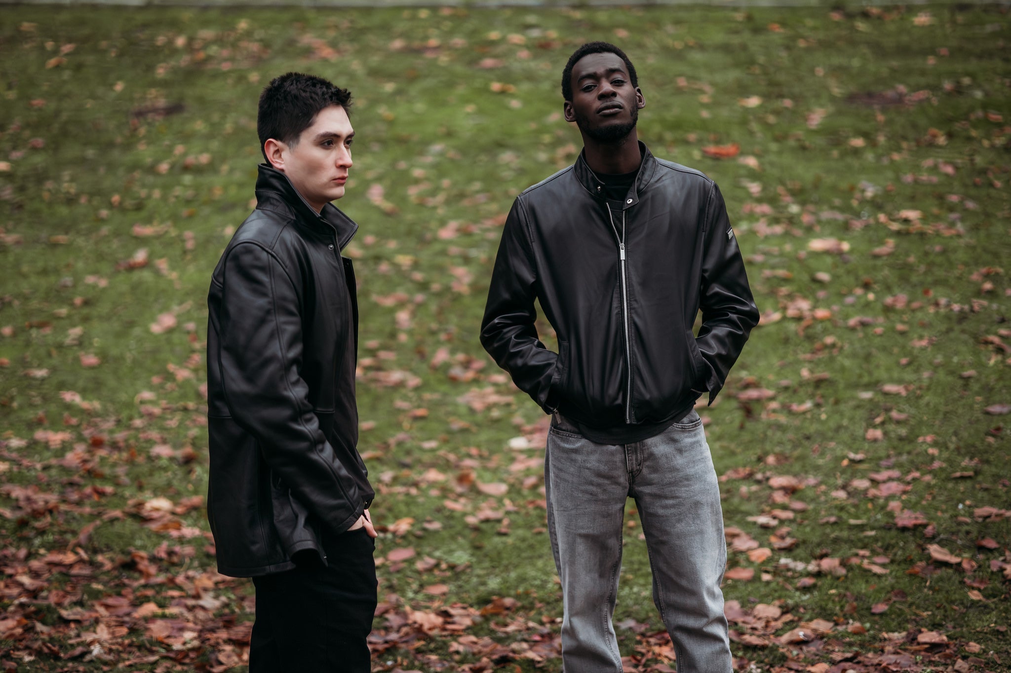 Two men standing wearing black Peroye leather jackets having a green grassy background.