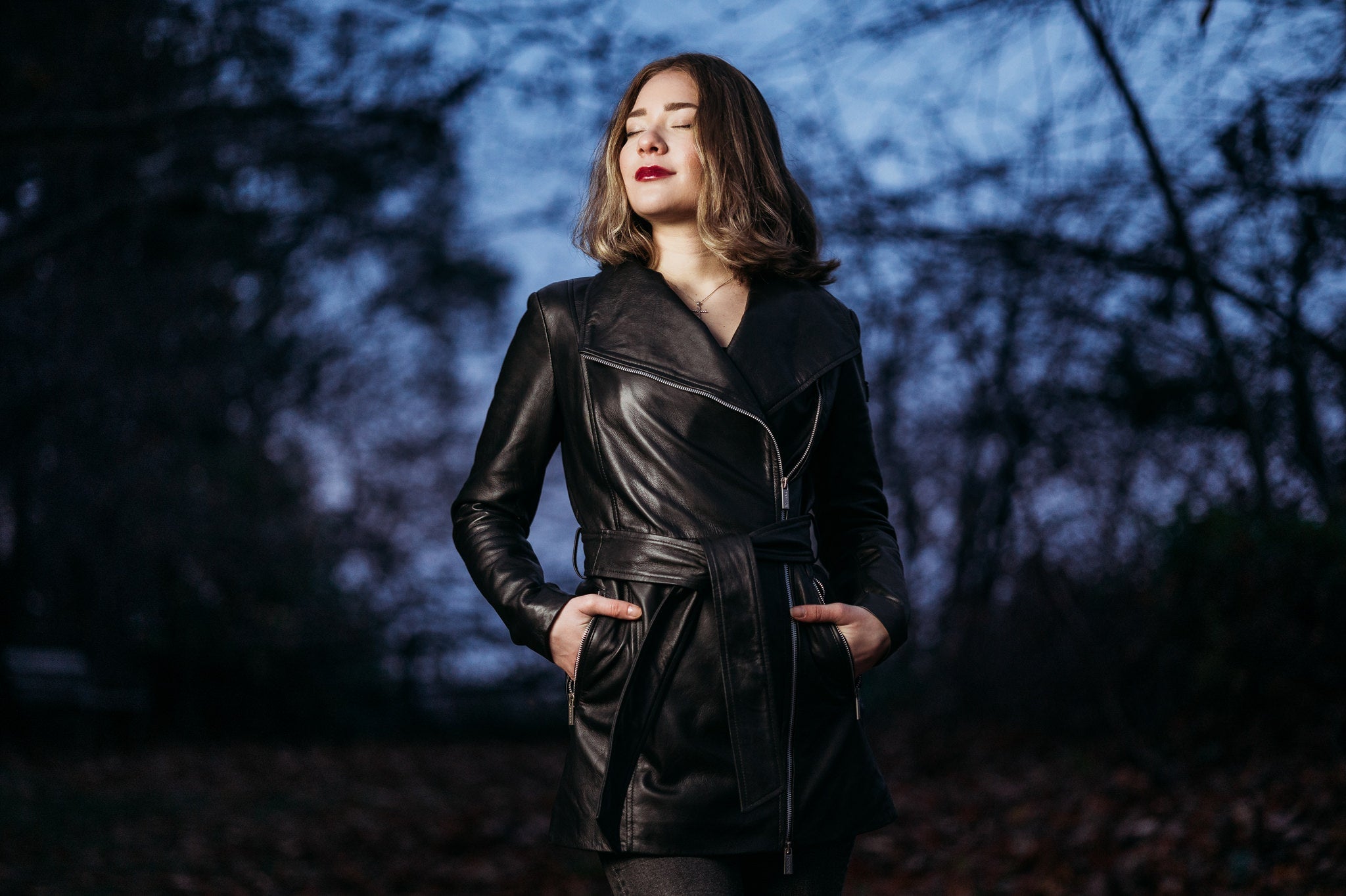 A woman standing wearing black Peroye leather jacket having a dark background with trees.