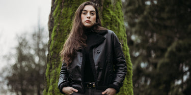 Peroye female model wearing black double breasted leather jacket with a lush green tree in her background.