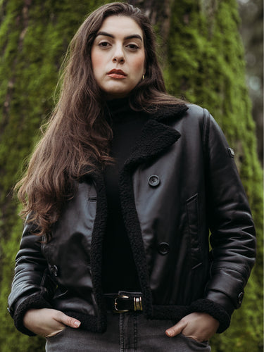 Peroye female model wearing black double breasted leather jacket with a lush green tree in her background.