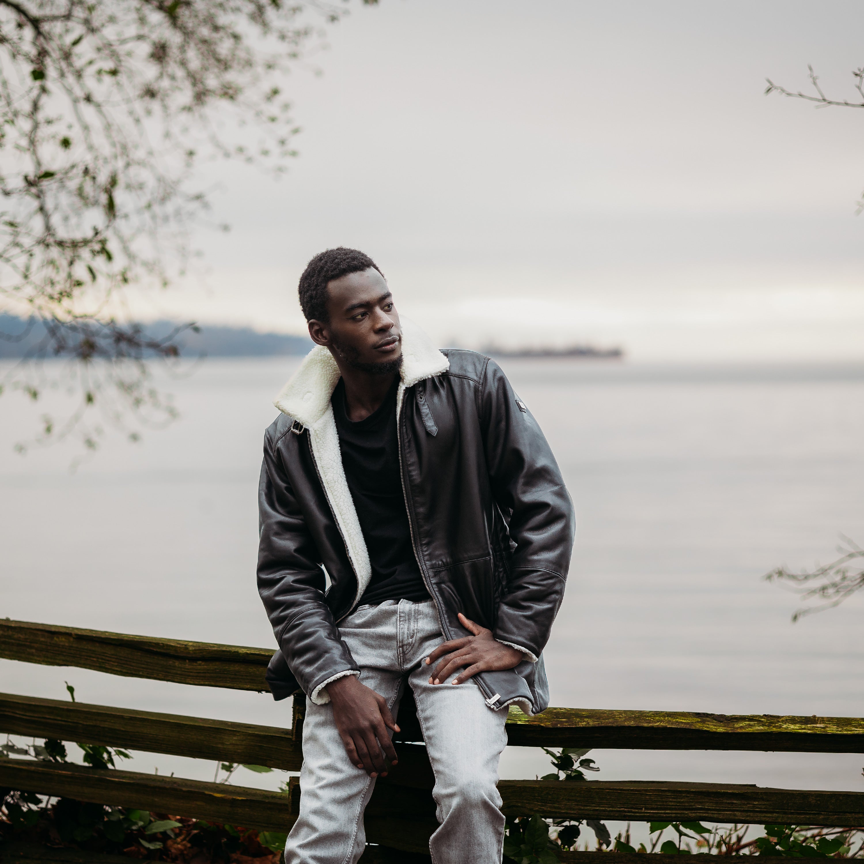 Peroye Male Model Wearing Black Leather Jacket With White Fur Lined. This is a trench coat style jacket. The model has an ocean in the background.