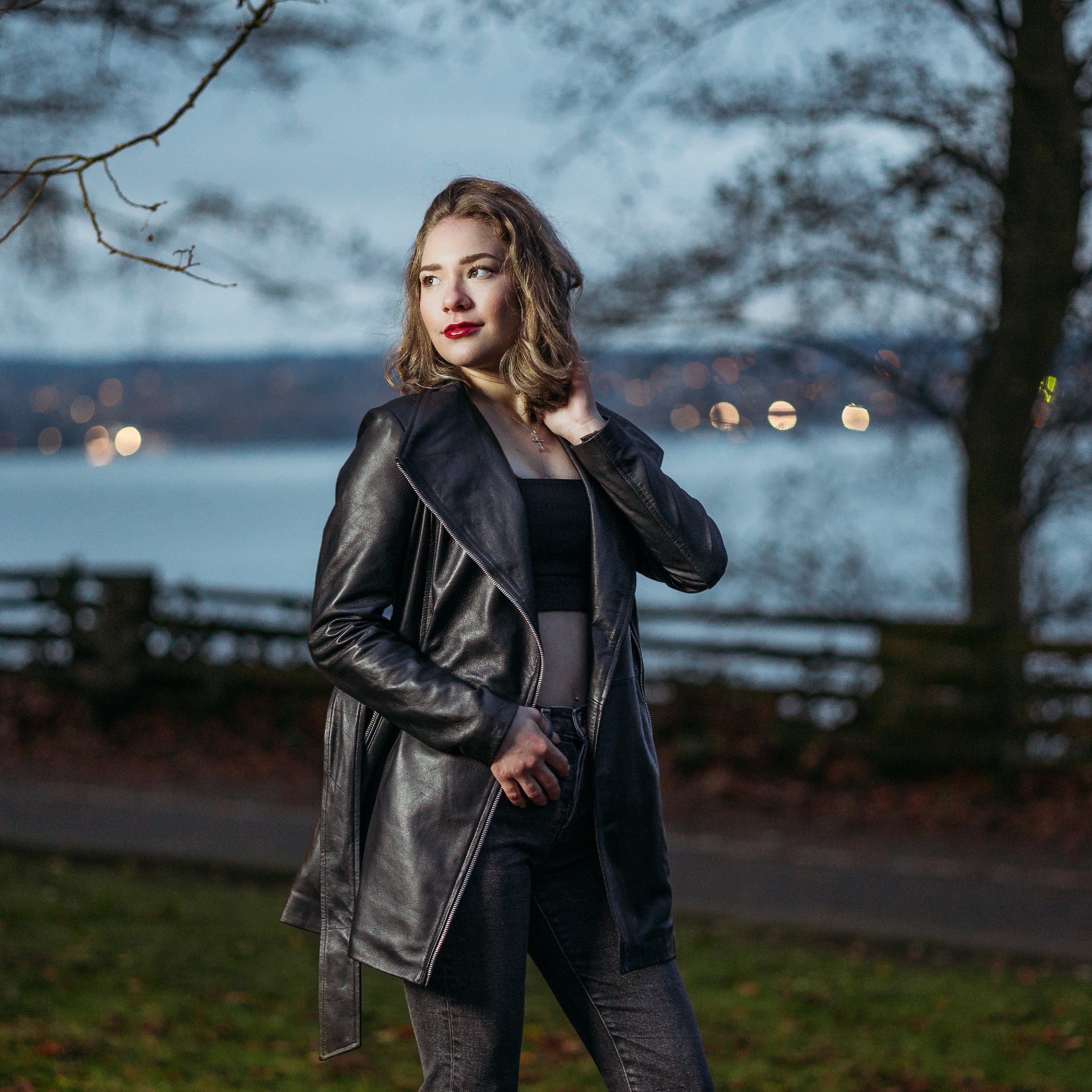 Peroye Female Model Wearing A Black Leather Jacket With Black Fur Lining. This is a trench coat style jacket. The model has a greenish background with trees.